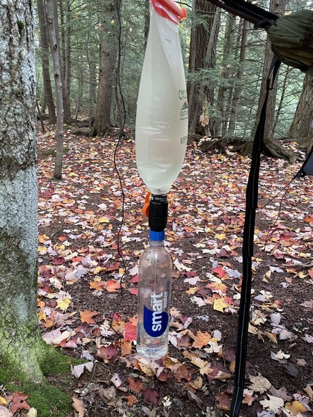 A pouch of captured water being gravity-fed through a Sawyer Squeeze into a drinking water bottle