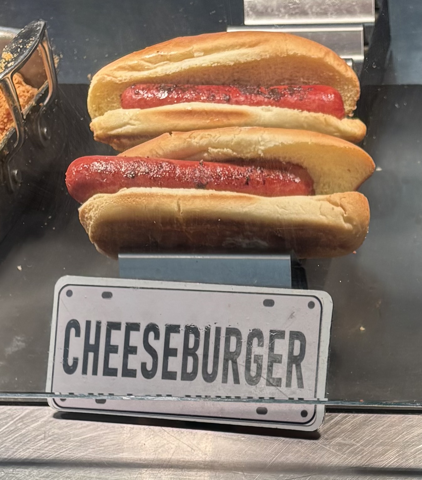 Grilled hot dogs in stale-looking buns behind a sign that reads “CHEESEBURGER”