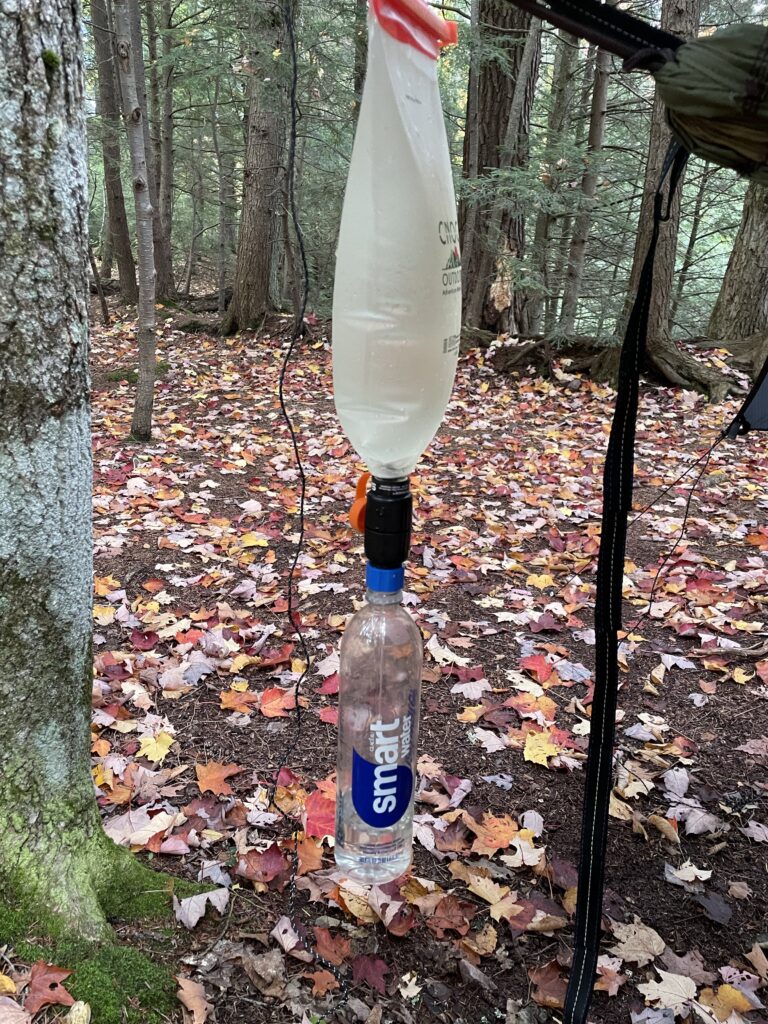 Little Carp River water being gravity-fed from a collapsible bladder through a Sawyer Squeeze Micro into a water bottle